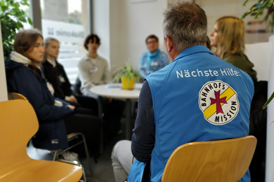 Das Bild zeigt den Raum der Ökumenischen Bahnhofsmission Dresden. Menschen sitzen an einem Tisch. Darunter ein Mitarbeiter der Bahnhofsmission mit entsprechender Weste. 