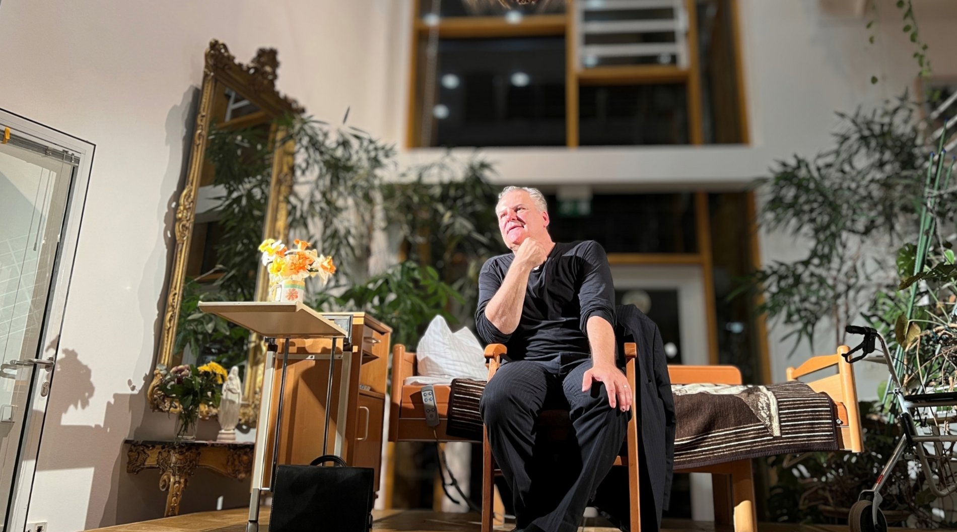 Schauspieler Tom Quaas sitzt im Altenpflegeheim Radebeul auf der Bühne. Hinter ihm steht ein Pflegebett. 