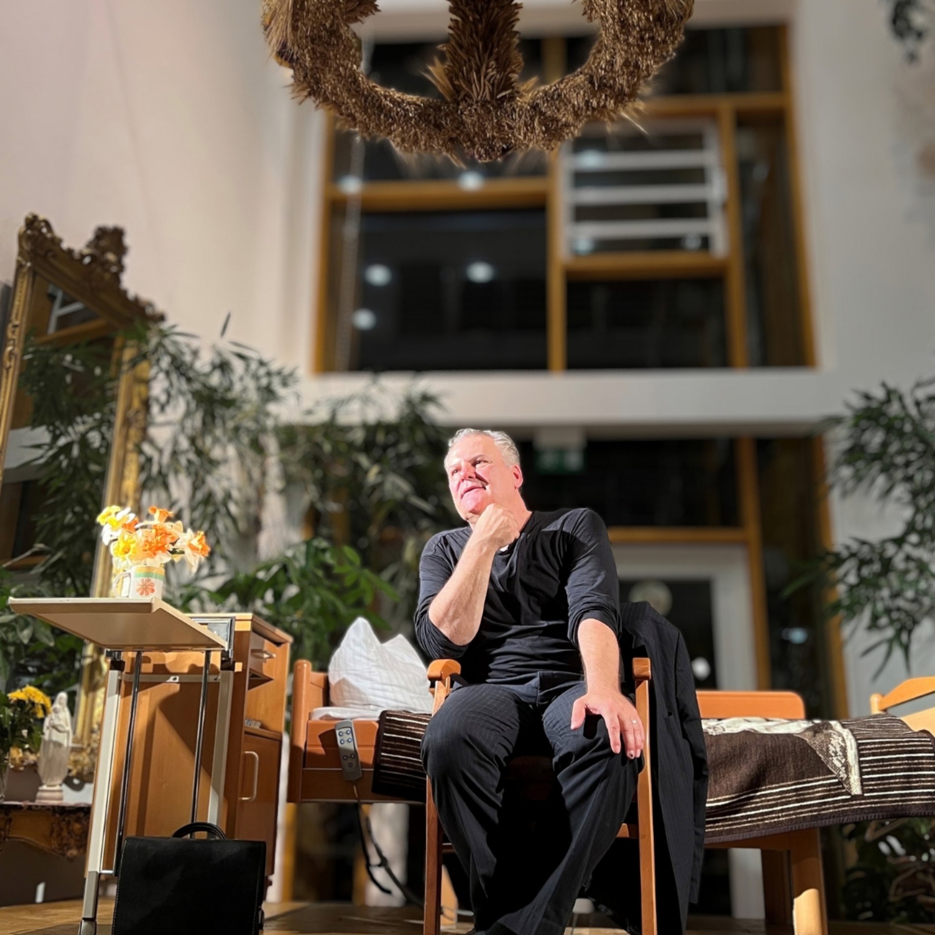 Schauspieler Tom Quaas sitzt im Altenpflegeheim Radebeul auf der Bühne. Hinter ihm steht ein Pflegebett. 