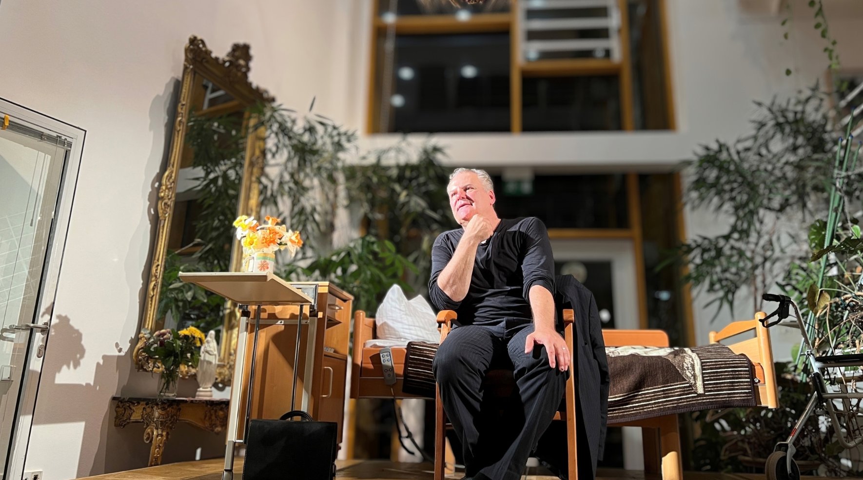 Schauspieler Tom Quaas sitzt im Altenpflegeheim Radebeul auf der Bühne. Hinter ihm steht ein Pflegebett. 