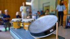 Auf diesem Bild sind im Vordergrund Percussions-Instrumente zu sehen. Im Hintergrund stehen Kinder und zwei Pädagoginnen im Halbkreis und klatschen zur Musik.
