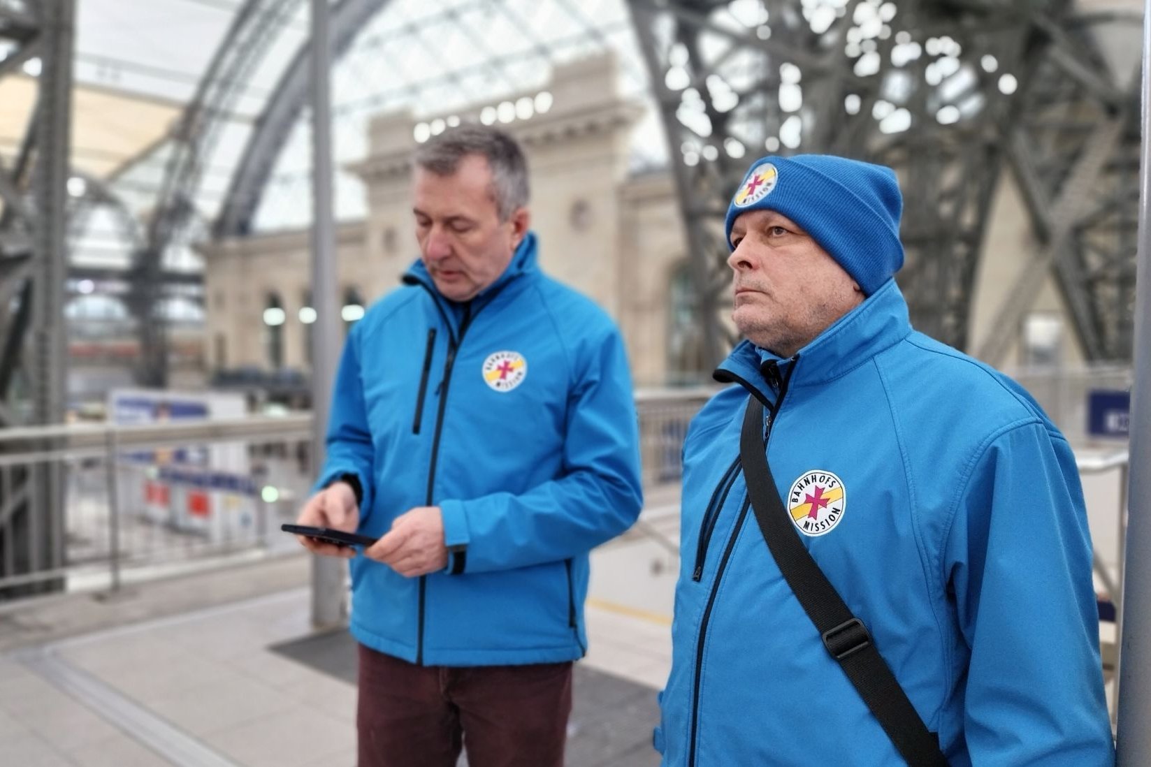 Das Bild zeigt Patrick Büttner, Koordinator der Bahnhofsmission Dresden, zusammen mit einem ehrenamtlichen Mitarbeiter. Die beiden sind an einem Bahnsteig am Dresdner Hauptbahnhof unterwegs.