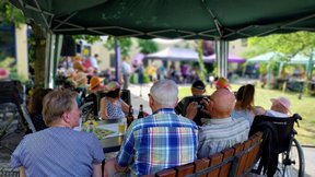 Bewohner:innen und Besucher:innen sitzen unter einem Pavillon am Tisch und lauschen der Andacht von Pfarrer Thomas Slesazeck.