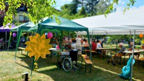 Dieses Bild zeigt den Garten der Friedenshöhe, auf dem mehrere Pavillons aufgebaut sind. Darunter stehen Biertischgarnituren, an denen Besucher:innen und Bewohner:innen sitzen. Alles ist bunt geschmückt, z.B. mit bunten Luftballons.