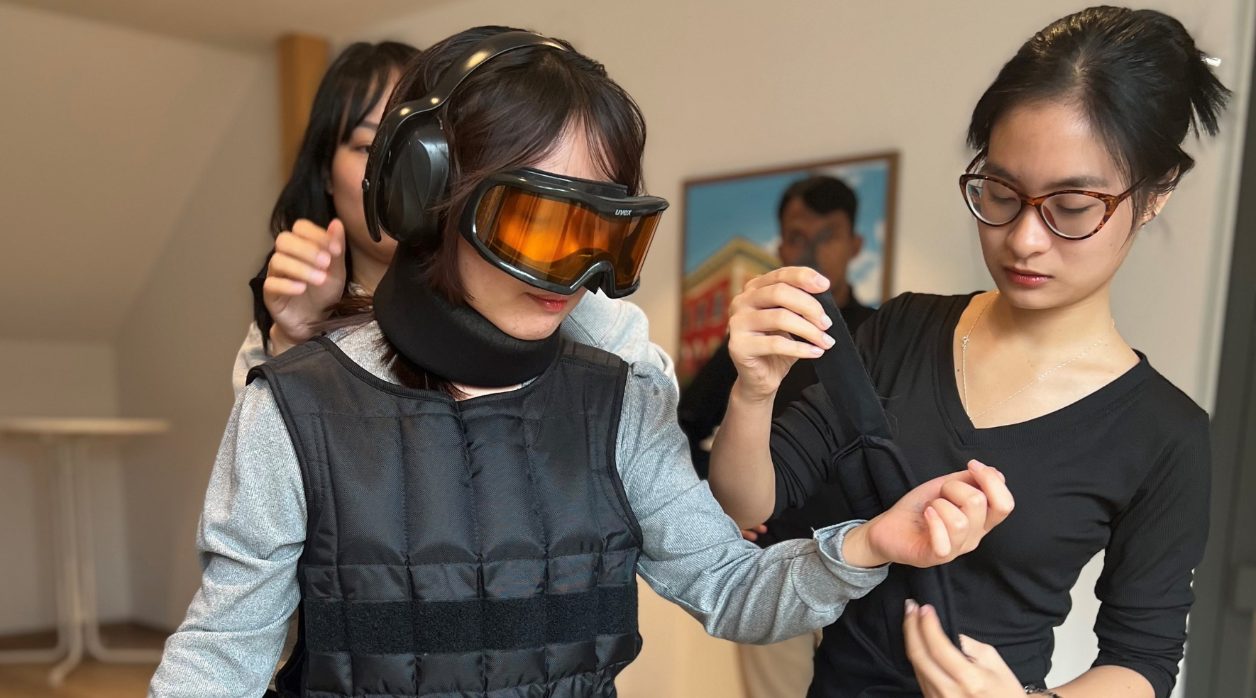 Eine junge Frau hilft einer zweiten Frau beim Anziehen eines sogenannten Alterssimulators. Er besteht unter anderem aus einer Art Ski-Brille und einem Gehörschutz. Auf dem Bild ist zu sehen, wie der Frau nun auch noch die dazugehörigen Gewichte um die Arme gelegt werden. 