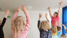Auch auf diesem Bild strecken die Yoga-Kinder ihre Arme unter Anleitung der Yoga-Lehrerin in die Luft.