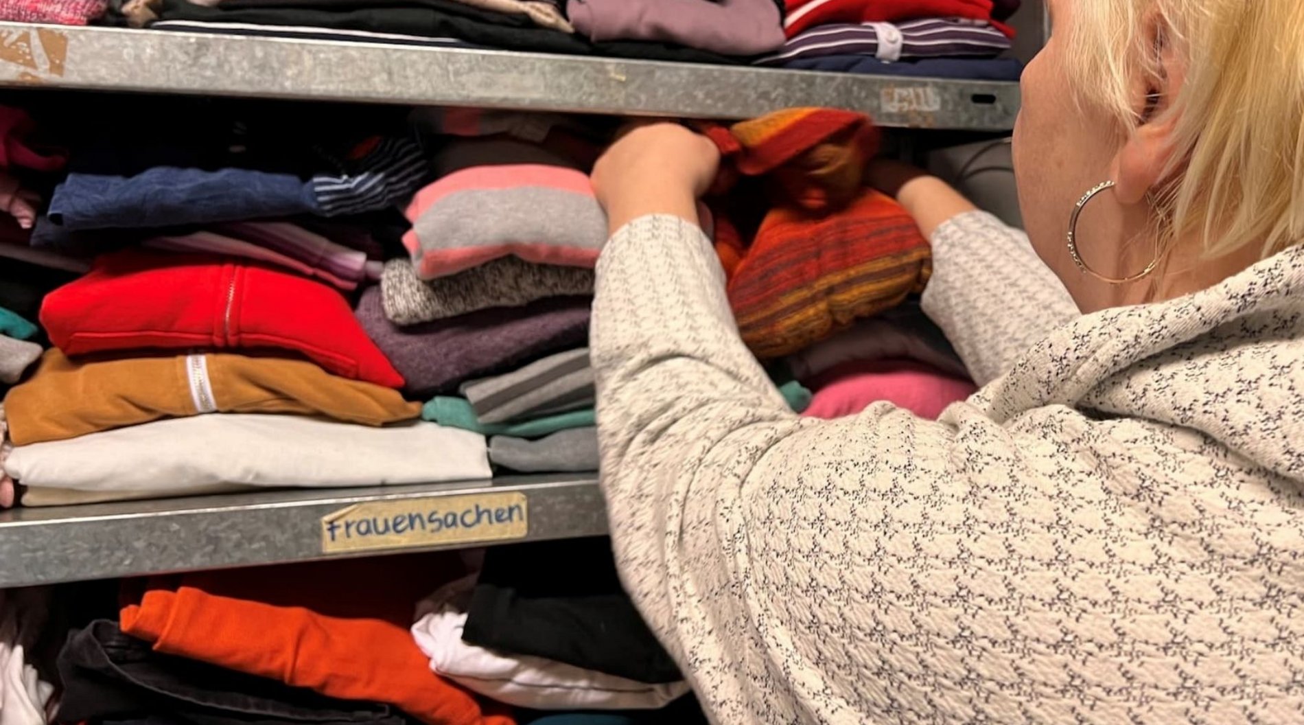 AUf diesem Bild ist eine Frau von hinten zu sehen, die Kleidung für Frauen aus einem Regal der Kleiderkammer nimmt.