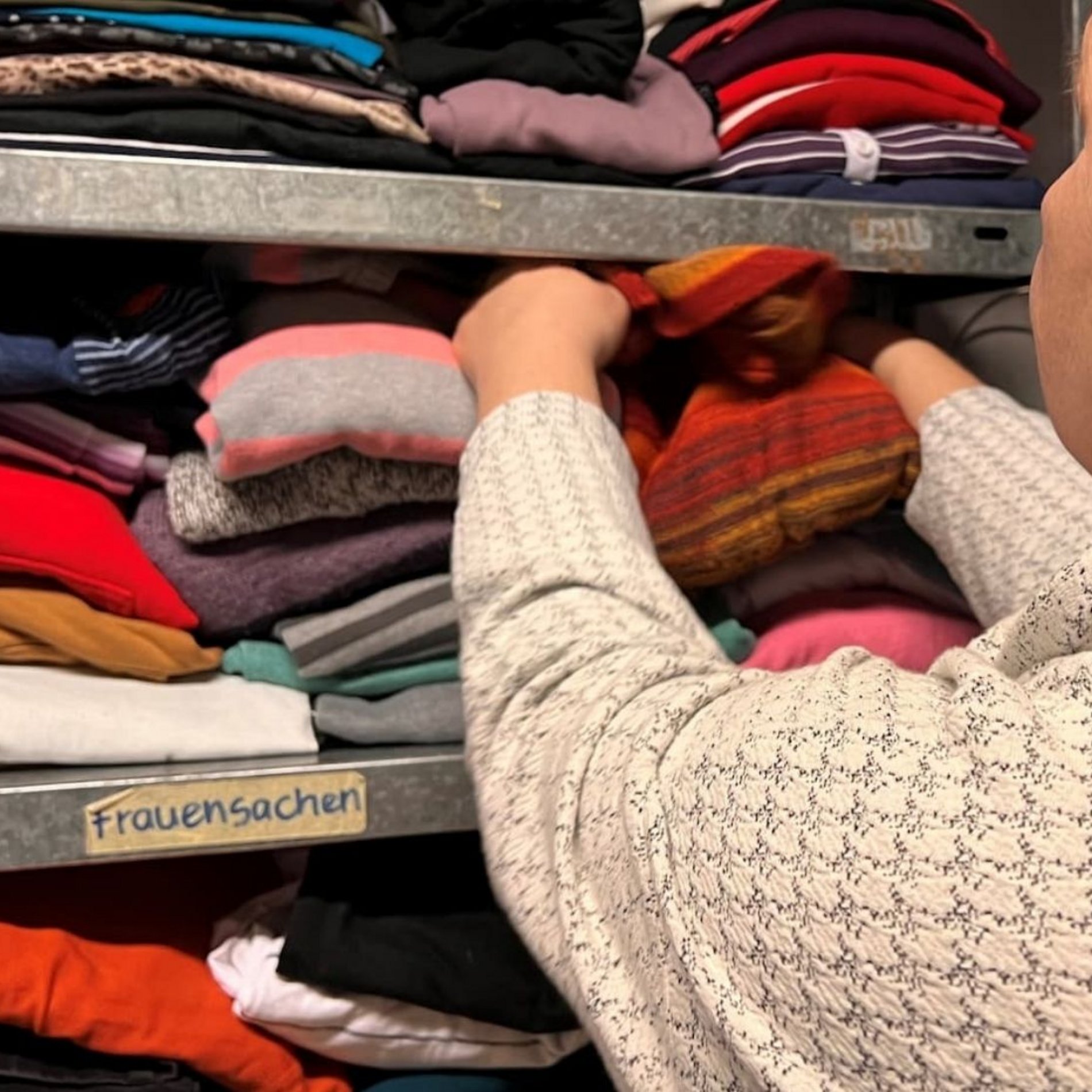 AUf diesem Bild ist eine Frau von hinten zu sehen, die Kleidung für Frauen aus einem Regal der Kleiderkammer nimmt.