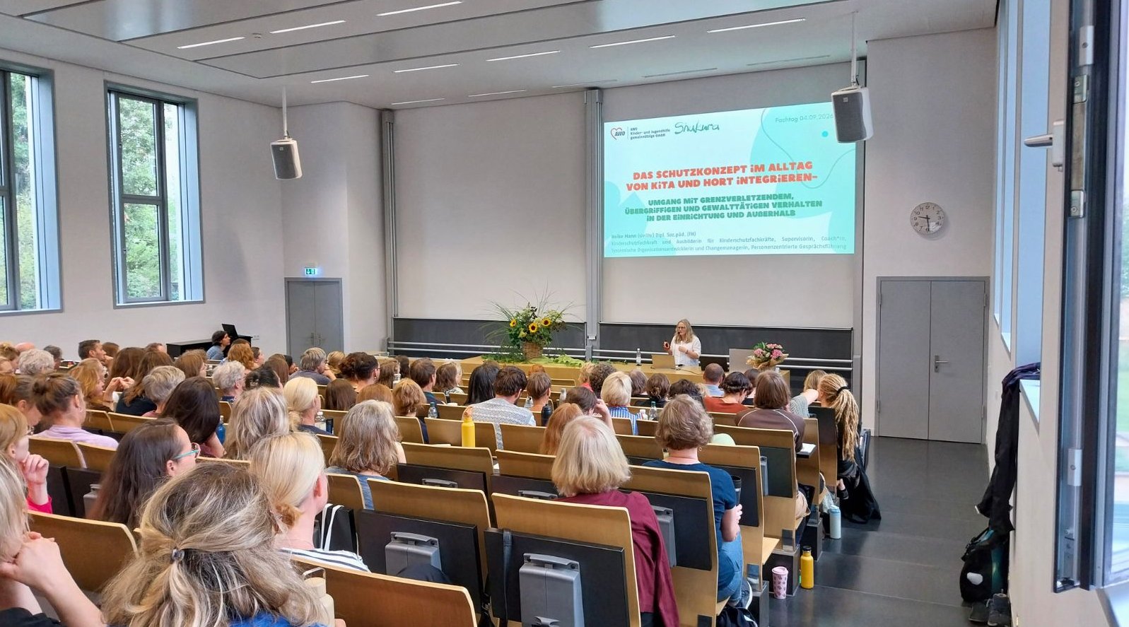 Dieses Bild zeigt den Hörsaal in der Evangelischen Hochschule Dresden, in dem der Abteilungsfachtag stattfand. Das Publikum ist von hinten zu sehen. Vorne steht Referentin Heike Mann während ihres ersten Impulsreferats.