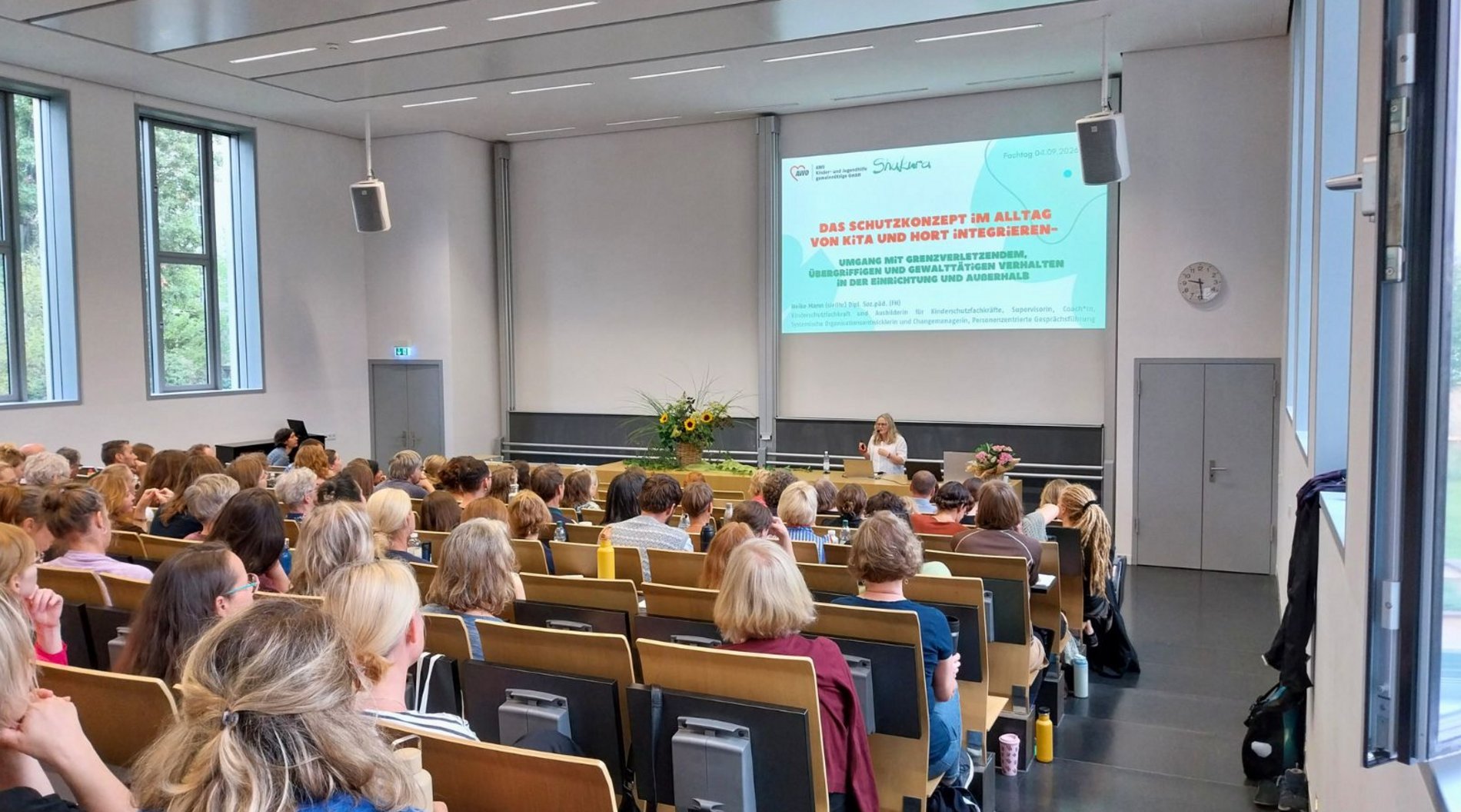 Dieses Bild zeigt den Hörsaal in der Evangelischen Hochschule Dresden, in dem der Abteilungsfachtag stattfand. Das Publikum ist von hinten zu sehen. Vorne steht Referentin Heike Mann während ihres ersten Impulsreferats.