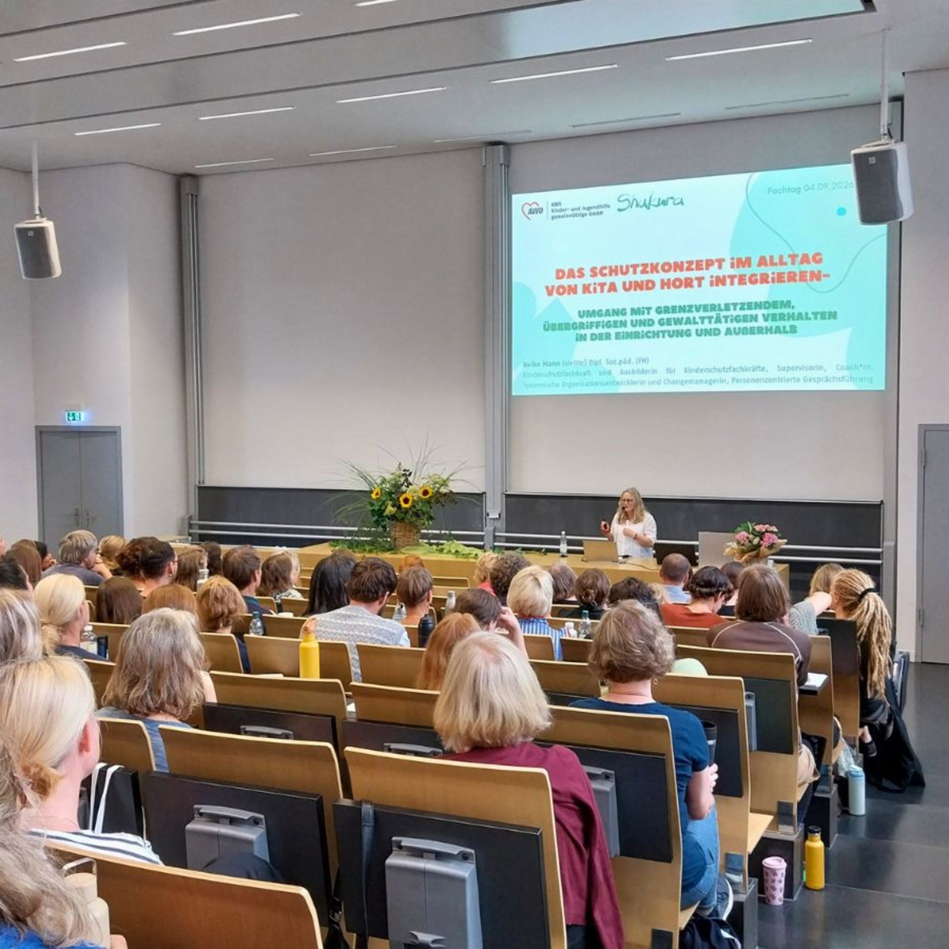 Dieses Bild zeigt den Hörsaal in der Evangelischen Hochschule Dresden, in dem der Abteilungsfachtag stattfand. Das Publikum ist von hinten zu sehen. Vorne steht Referentin Heike Mann während ihres ersten Impulsreferats.