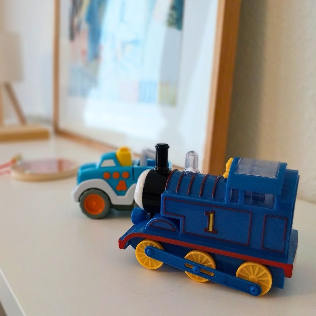 Auf dem Bild ist ein Regal zu sehen, auf dem eine Lampe, ein Bild sowie Spielzeugfahrzeuge stehen. Das Bild wurde im Wohnzimmer der Wohngruppe Komet aufgenommen.