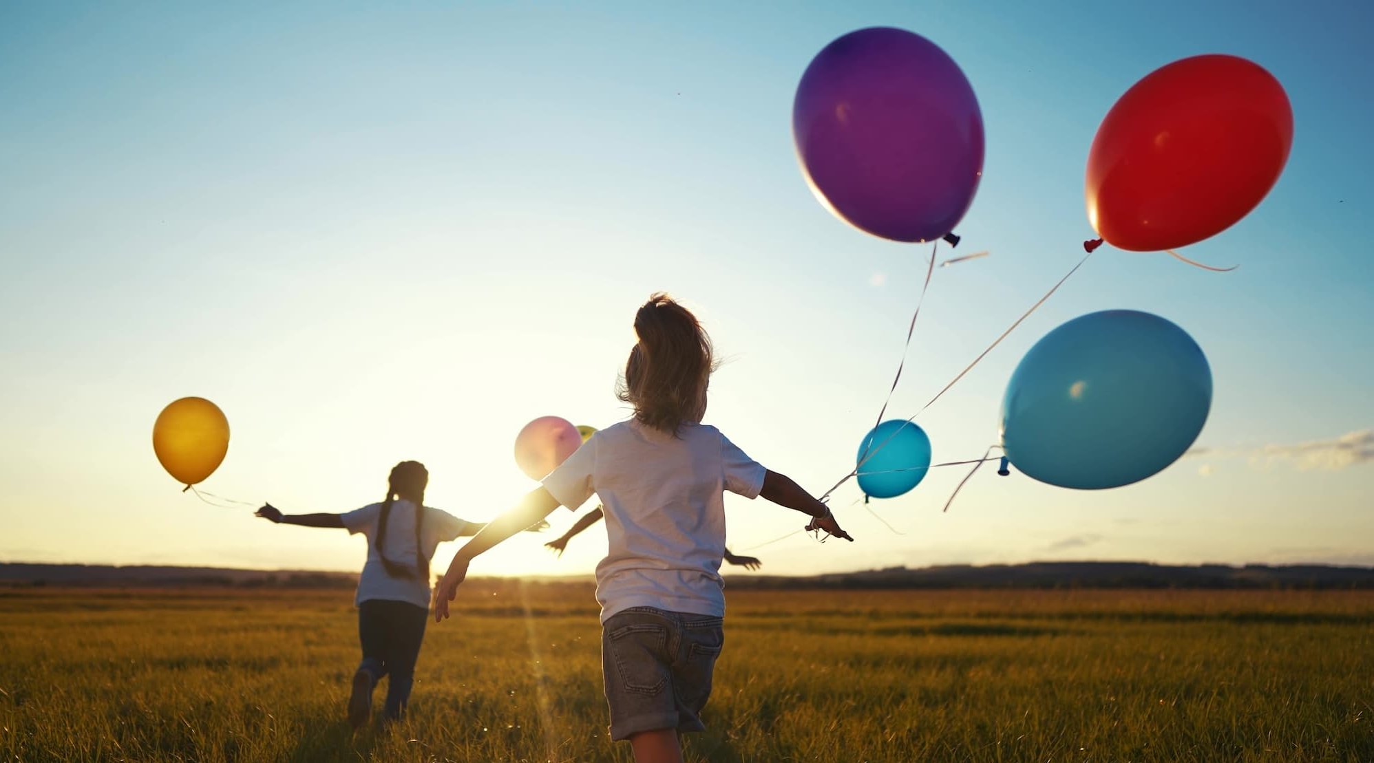 Kinder laufen auf einer Wiese in den Sonnenuntergang, in der Hand bunte Lufftballons