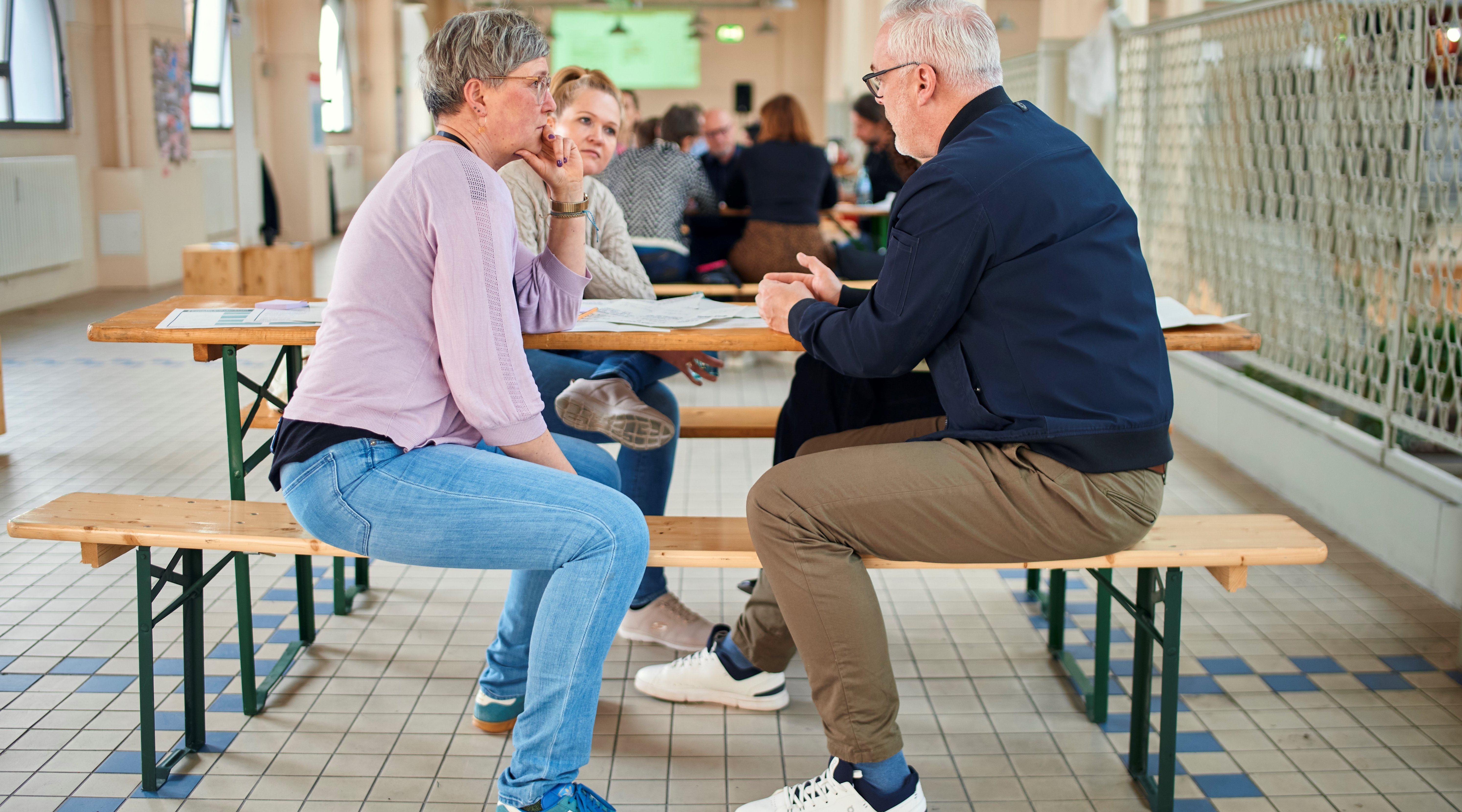 Dieses Bild zeigt eine Beratungssituation in der "Malle". Drei Menschen sitzen zusammen an einem Tisch in der Markthalle und reden miteinander.