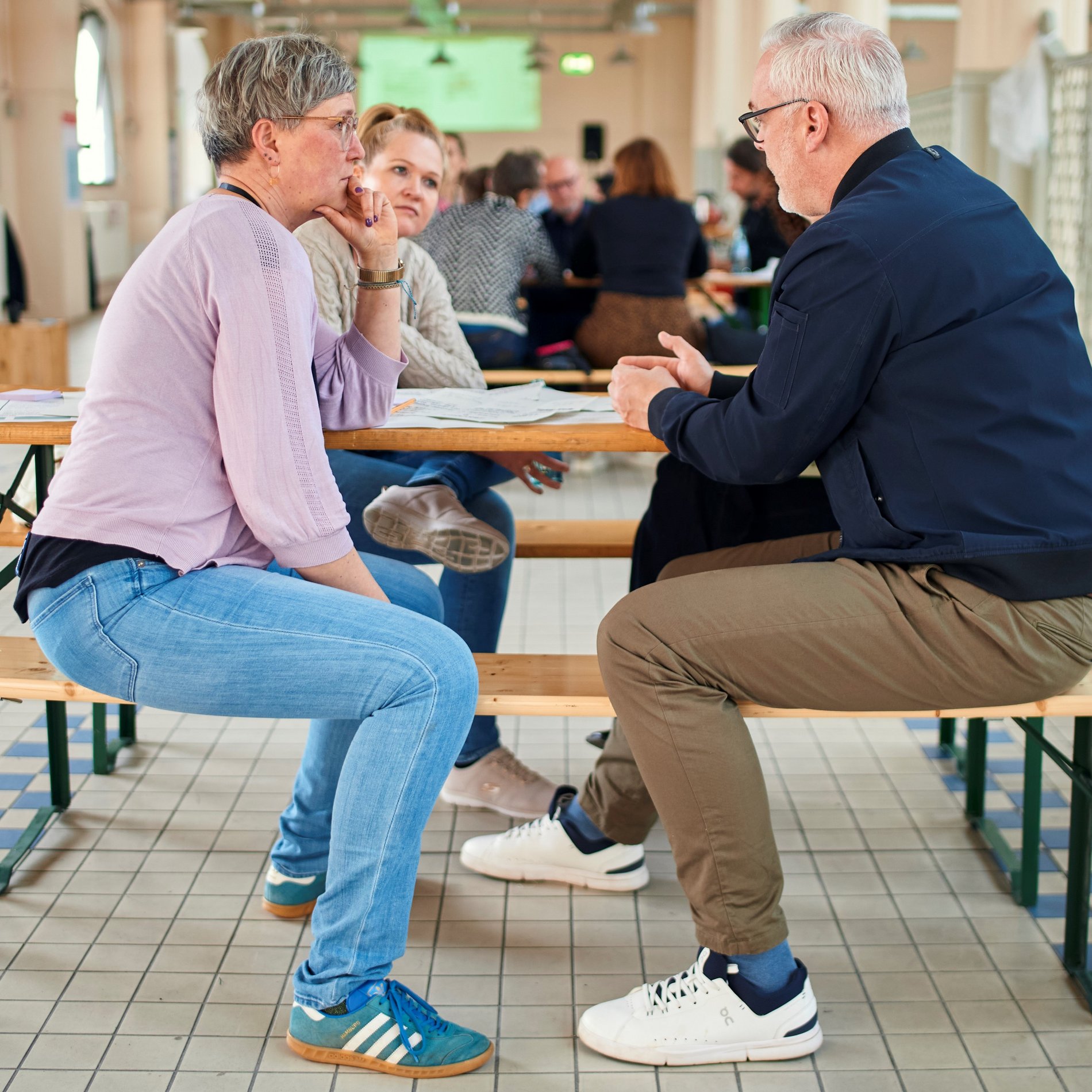Dieses Bild zeigt eine Beratungssituation in der "Malle". Drei Menschen sitzen zusammen an einem Tisch in der Markthalle und reden miteinander.